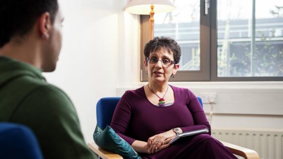 A male and a female sat on chairs facing towards each other in a room, the female has a clipboard and is listening to the male.
